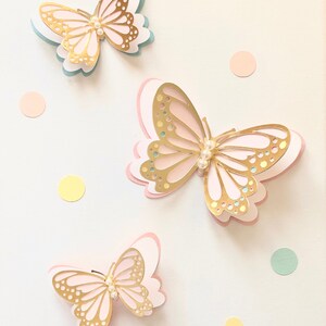 Puede incluir: Tres mariposas doradas con detalles de perlas sobre un fondo blanco con puntos de colores pastel.
