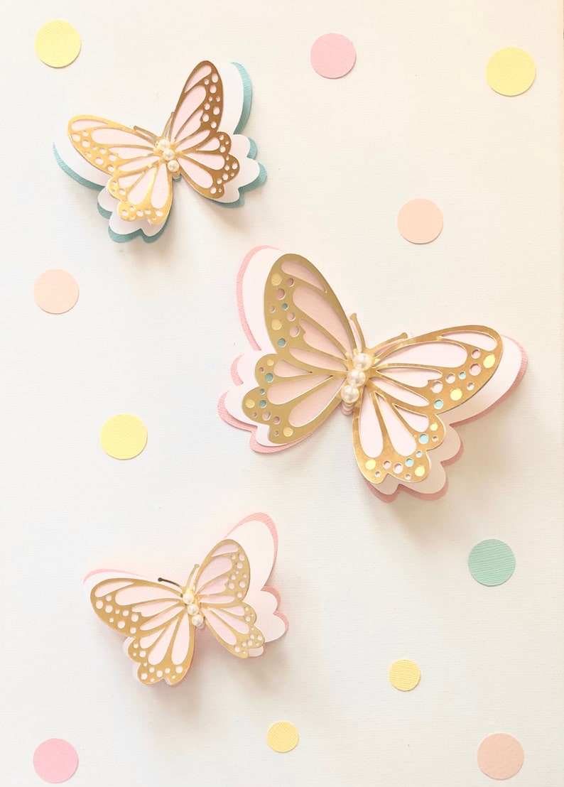 Puede incluir: Tres mariposas doradas con detalles de perlas sobre un fondo blanco con puntos de colores pastel.
