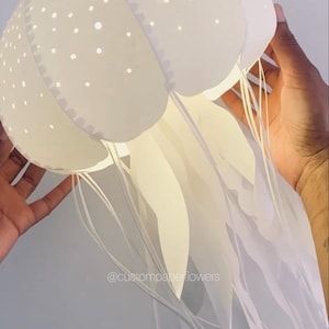 Puede incluir: Una lámpara de medusa de papel blanco con una cúpula redondeada con patrones de puntos perforados y largos tentáculos de papel fluidos. La estructura está iluminada desde el interior, emitiendo un brillo suave. El texto @custompaperflowers es visible.