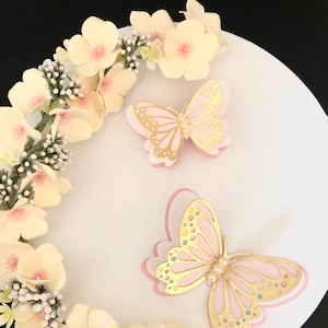 Puede incluir: Dos delicadas mariposas rosas y doradas con detalles de perlas sobre un pastel blanco decorado con una corona de flores de color amarillo p&aacute;lido.