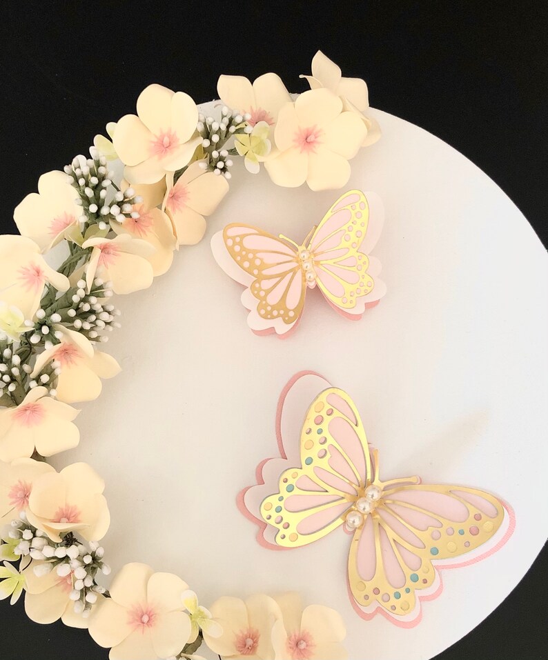 Puede incluir: Dos delicadas mariposas rosas y doradas con detalles de perlas sobre un pastel blanco decorado con una corona de flores de color amarillo p&aacute;lido.