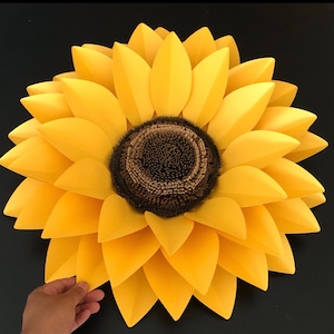May include: A large, paper sunflower with yellow petals and a brown center. The sunflower is made of multiple layers of paper petals.
