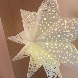 Peut inclure: Un abat-jour suspendu en forme d'étoile blanche à plusieurs branches, doté de motifs complexes découpés au laser, composés de formes circulaires et tourbillonnantes. La structure en papier plié laisse passer la lumière à travers les découpes.