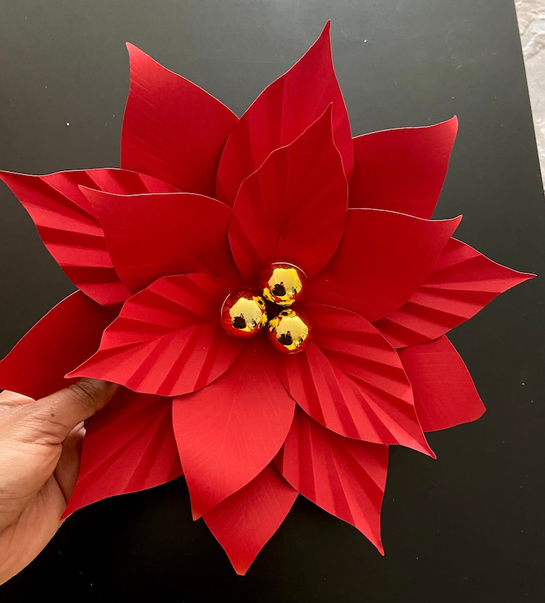May include: A red paper poinsettia flower with three gold ornaments in the center.