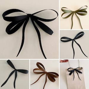 May include: A collection of decorative bows made from smooth, flat ribbon material. The bows are shown in various solid colours including black, olive green, and brown. Each bow features two large loops and two long, trailing ribbon ends.