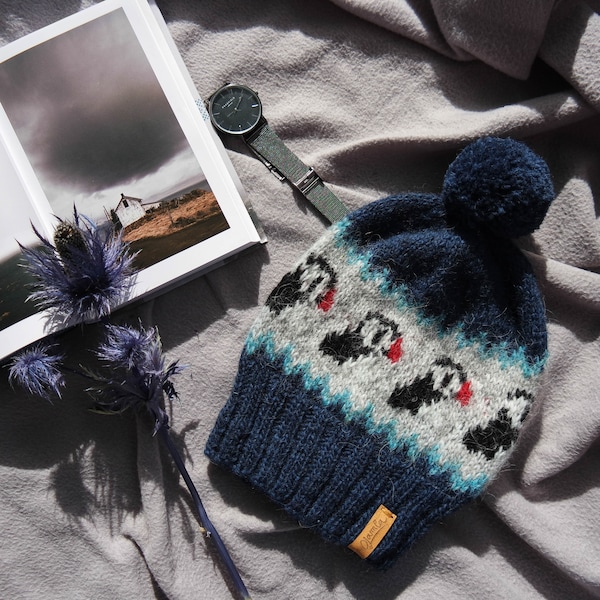 May include: A navy blue knit beanie with a pom-pom on top, featuring a light gray band with a repeating pattern of black and white puffins with red beaks. The beanie has a ribbed cuff and a small tan leather tag with the brand name Jamla.