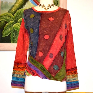 May include: A long-sleeve shirt featuring a patchwork design with diagonal panels in red, blue, and green. The garment includes circular fabric appliqués, exposed seams, and horizontal bands of floral and geometric patterns on the cuffs.