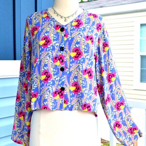 Pode incluir: Uma blusa de manga comprida com botões numa base azul vibrante com um padrão repetitivo de flores semelhantes a amores-perfeitos em rosa e amarelo e folhagem clara. A peça tem um decote em V e quatro botões circulares pretos na frente.