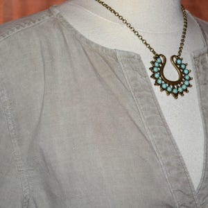 May include: A light gray shirt with a V-neck collar is displayed on a mannequin. A gold-colored necklace with a horseshoe pendant and turquoise-colored stones is worn over the shirt. The necklace is the focal point of the image.