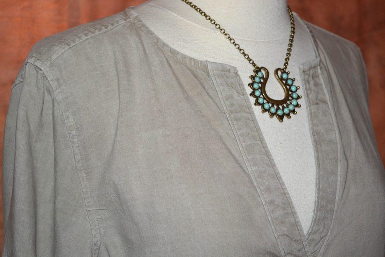 May include: A light gray shirt with a V-neck collar is displayed on a mannequin. A gold-colored necklace with a horseshoe pendant and turquoise-colored stones is worn over the shirt. The necklace is the focal point of the image.