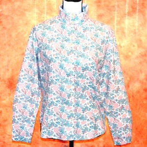 May include: A long-sleeved button-down shirt featuring a high collar and a repeating floral pattern in shades of muted pink, sage green, and cream. The garment has a central vertical row of small buttons and a structured fit with long sleeves.