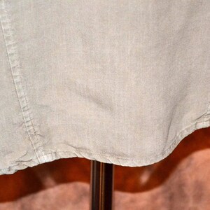 May include: Close-up of a light beige linen shirt. The fabric has a textured appearance with visible seams and a curved hemline. The shirt is displayed on a stand, with the bottom portion of the garment visible.