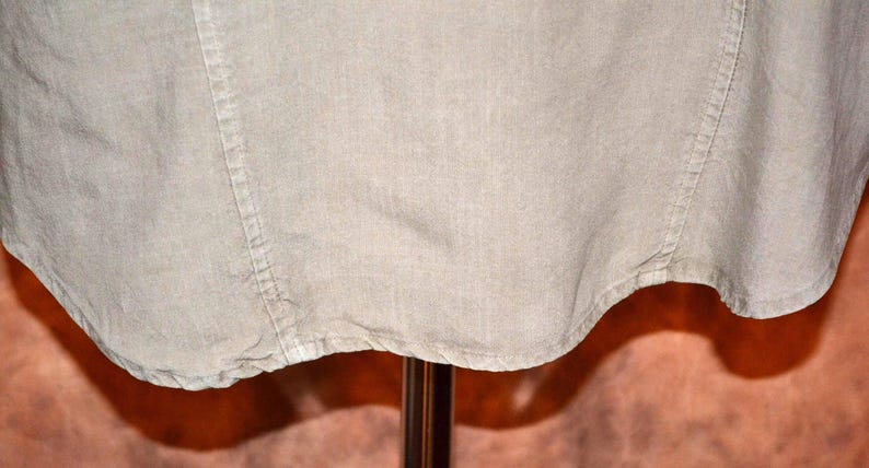 May include: Close-up of a light beige linen shirt. The fabric has a textured appearance with visible seams and a curved hemline. The shirt is displayed on a stand, with the bottom portion of the garment visible.
