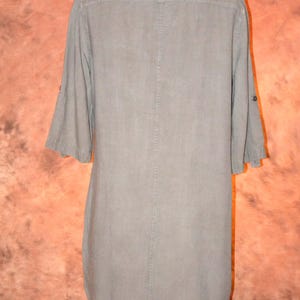 May include: A light olive green tunic dress with a button-down front and three-quarter sleeves. The dress is displayed on a mannequin, showcasing its simple, yet elegant design. The fabric appears to be lightweight.
