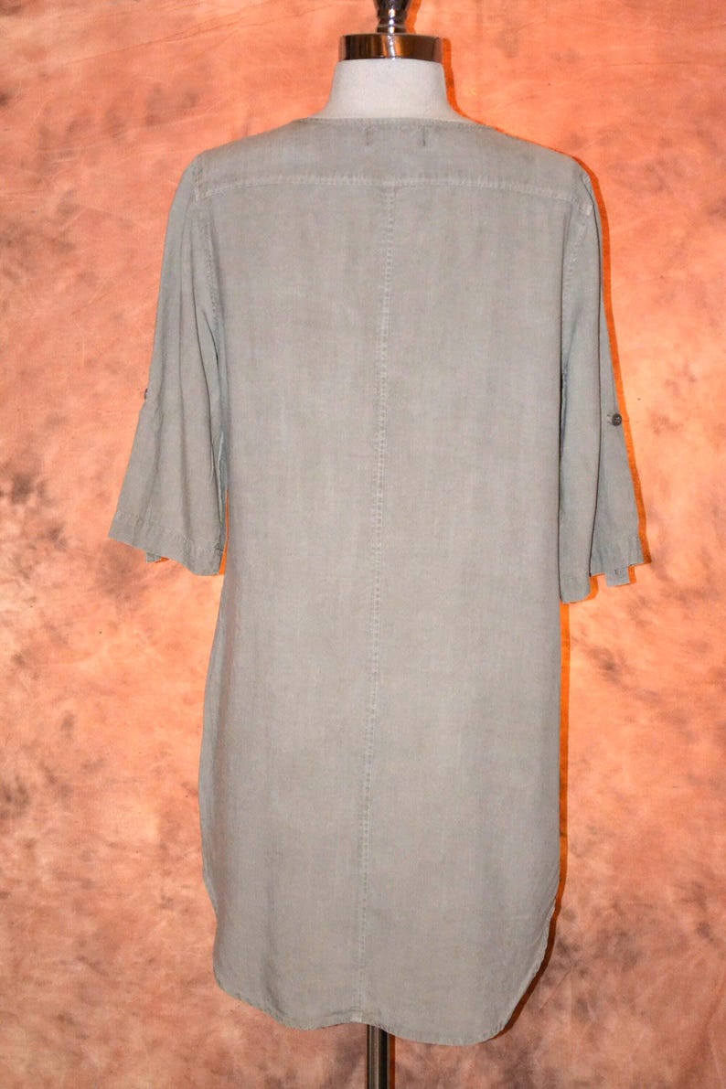 May include: A light olive green tunic dress with a button-down front and three-quarter sleeves. The dress is displayed on a mannequin, showcasing its simple, yet elegant design. The fabric appears to be lightweight.
