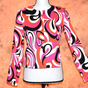 May include: A vibrant, patterned jacket in a retro style. The jacket features a bold design with swirls of pink, orange, black, and white. It has long sleeves, a single-button closure, and a round neckline. The jacket is displayed on a mannequin.