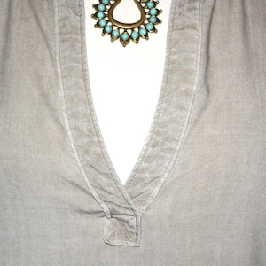 May include: Close-up of a light gray shirt with a V-neck collar. A gold-toned necklace with turquoise-colored stones is visible at the neckline. The shirt has long sleeves and a textured appearance.