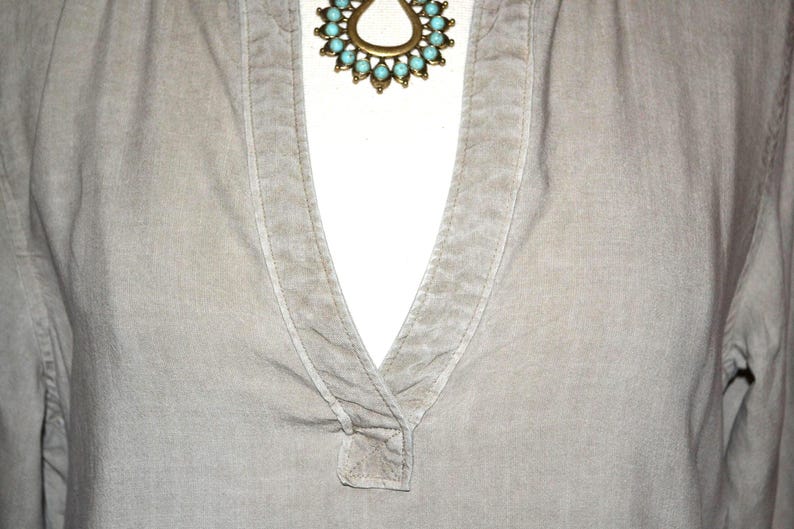 May include: Close-up of a light gray shirt with a V-neck collar. A gold-toned necklace with turquoise-colored stones is visible at the neckline. The shirt has long sleeves and a textured appearance.