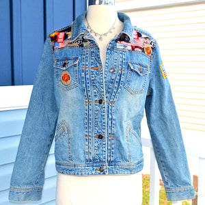 May include: A light blue denim jacket featuring a collared neckline, long sleeves, and a button-down front. The upper chest area is decorated with various colourful patches and fabric appliques, while a small American flag patch is on the left sleeve.