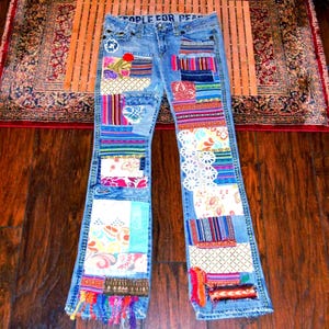 Pode incluir: Jeans de ganga azul claro com designs de patchwork coloridos. Os jeans apresentam vários padrões de tecido, incluindo florais, listrados e paisley, com as palavras "PEOPLE FOR PEACE" na parte superior. Os jeans têm uma perna flare e são decorados com franjas.