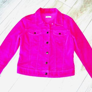 Pode incluir: Um casaco de veludo cotelê em rosa choque com gola clássica, mangas compridas e fecho frontal de botões. A peça inclui dois bolsos no peito com abas abotoadas e uma cintura estruturada, apresentando um acabamento de tecido texturizado.