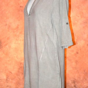 May include: A light gray tunic dress with a V-neckline and three-quarter sleeves. The dress features a button detail on the sleeves and a decorative necklace. The dress is displayed on a mannequin.