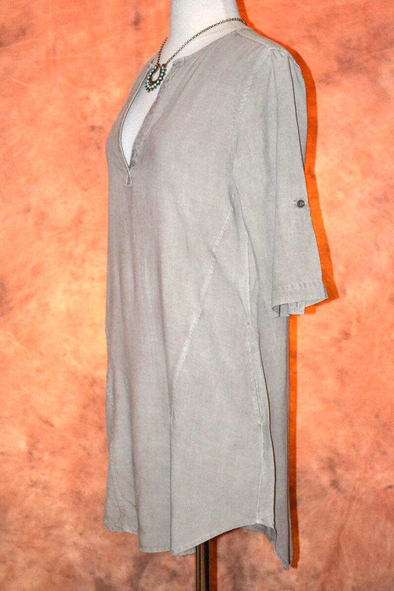 May include: A light gray tunic dress with a V-neckline and three-quarter sleeves. The dress features a button detail on the sleeves and a decorative necklace. The dress is displayed on a mannequin.