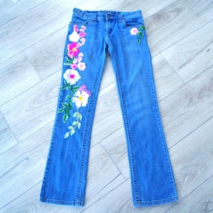 May include: Medium-wash blue denim jeans featuring a floral embroidery pattern. The design includes white, pink, and magenta flowers with green leaves cascading down the right leg and a smaller floral accent near the left front pocket.