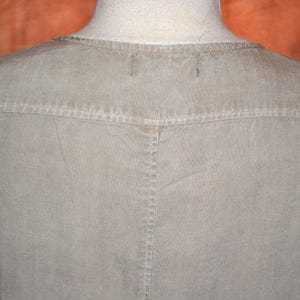 May include: Close-up of a light olive-green shirt. The shirt features a crew neck and a yoke detail across the shoulders. The stitching is visible, and the fabric appears to be a soft, woven material. The shirt is displayed on a mannequin.