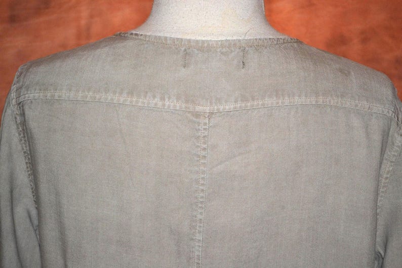 May include: Close-up of a light olive-green shirt. The shirt features a crew neck and a yoke detail across the shoulders. The stitching is visible, and the fabric appears to be a soft, woven material. The shirt is displayed on a mannequin.