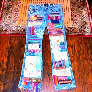 May include: Light blue denim jeans featuring a patchwork design with various colourful fabric patterns, lace inserts, and frayed hems. The waistband interior displays the text "PEOPLE FOR PEACE" in white lettering against a dark blue background.