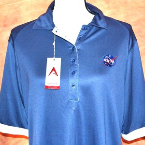 Puede incluir: Un polo de manga corta azul oscuro con ribete blanco en el cuello y puños blancos. La camisa presenta un logotipo circular de la NASA bordado en el pecho izquierdo y una etiqueta de marca rectangular blanca unida a la tapeta de botones.