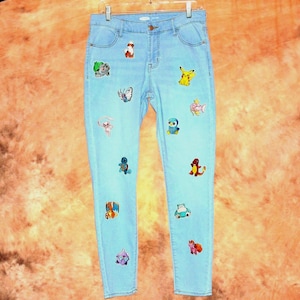 May include: Light blue denim jeans adorned with colorful embroidered patches of various cartoon characters. The jeans have a classic five-pocket design and are hanging on a metal hanger. The patches are of different sizes and shapes.