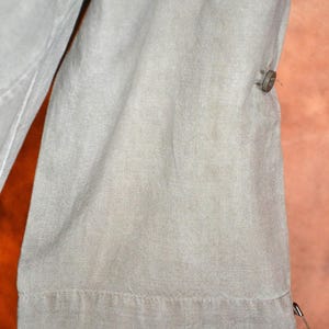May include: Close-up of a section of light green fabric, possibly a garment. The fabric has a textured appearance with visible stitching along the hem. A button and a safety pin are attached to the fabric.