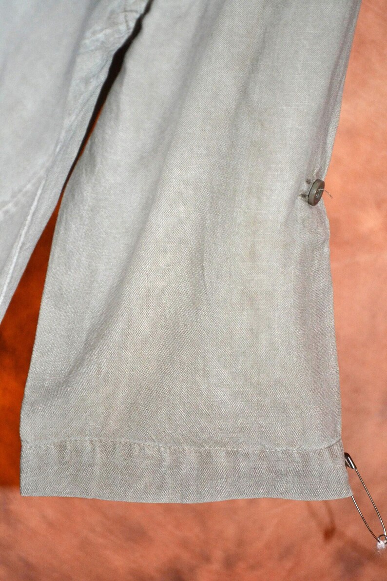 May include: Close-up of a section of light green fabric, possibly a garment. The fabric has a textured appearance with visible stitching along the hem. A button and a safety pin are attached to the fabric.