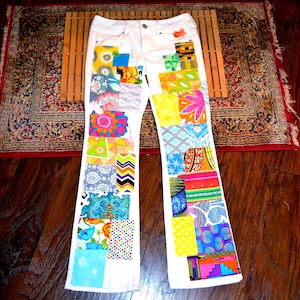 Könnte beinhalten: Weiße Jeans mit bunten Patchwork-Designs. Die Patches zeigen verschiedene Muster und Farben, darunter Blumen-, geometrische und abstrakte Designs. Die Jeans liegen flach auf einer Holzoberfläche mit einem gemusterten Teppich im Hintergrund.
