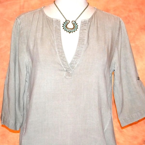 May include: A light gray tunic-style shirt with a V-neck and three-quarter sleeves. The shirt has a relaxed fit and a button detail on the sleeves. A turquoise and bronze necklace is worn with the shirt.