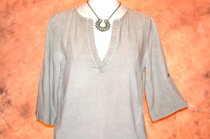 May include: A light gray tunic-style shirt with a V-neck and three-quarter sleeves. The shirt has a relaxed fit and a button detail on the sleeves. A turquoise and bronze necklace is worn with the shirt.