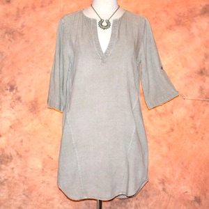 May include: A light gray tunic-style top with a V-neck and three-quarter sleeves. The top has a relaxed fit and a curved hemline. A dark metal necklace with a pendant is visible. The fabric appears to be a lightweight linen or cotton blend.