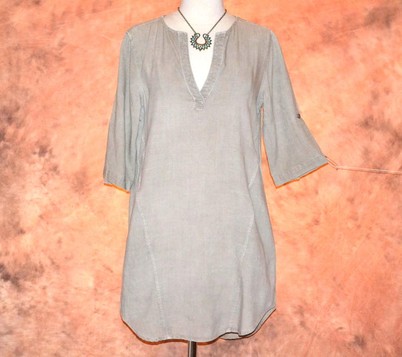May include: A light gray tunic-style top with a V-neck and three-quarter sleeves. The top has a relaxed fit and a curved hemline. A dark metal necklace with a pendant is visible. The fabric appears to be a lightweight linen or cotton blend.