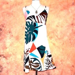 May include: A sleeveless white dress featuring a bold, multicolored graphic print of large tropical leaves in shades of dark green, brown, and blue, accented by geometric shapes in orange and pink, with a V-neckline and thin spaghetti straps.