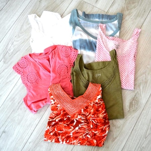 May include: A collection of six sleeveless tops featuring a variety of designs including a solid white top, a blue and white tie-dye top, a pink patterned top, a bright pink top with eyelet details, an olive green top, and a red floral print top.