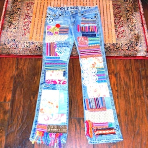 Può includere: Jeans in denim azzurro con un design patchwork caratterizzato da vari motivi in tessuto colorato, sovrapposizioni in pizzo e dettagli ricamati. L'interno della cintura riporta la scritta PEOPLE FOR PEACE. Gli orli delle gambe presentano frange.