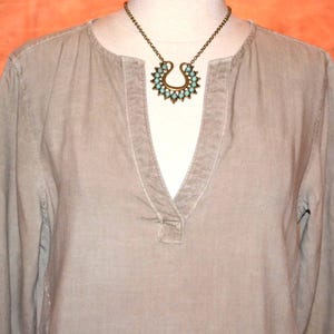 May include: A light olive green long-sleeve shirt with a V-neck collar. A bronze-colored necklace with a turquoise-colored pendant is displayed on the shirt. The pendant has a horseshoe shape with turquoise-colored stones.