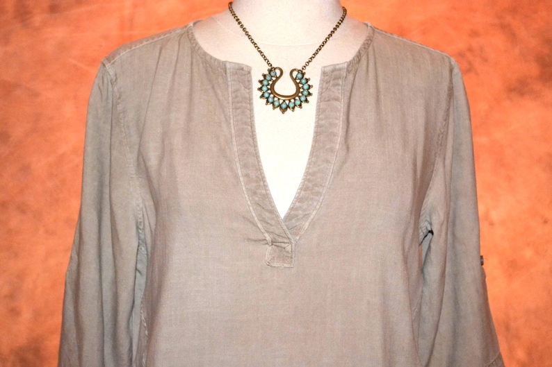 May include: A light olive green long-sleeve shirt with a V-neck collar. A bronze-colored necklace with a turquoise-colored pendant is displayed on the shirt. The pendant has a horseshoe shape with turquoise-colored stones.