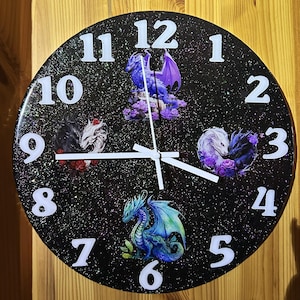Puede incluir: Reloj de pared redondo negro con números y manecillas blancas. La esfera del reloj presenta cuatro ilustraciones de dragones en tonos de azul, morado y blanco. El fondo es negro moteado con purpurina.