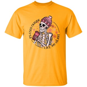 May include: Mustard yellow t-shirt featuring a skeleton wearing a pink beanie with hearts, holding a red cup. The graphic includes the text "IT'S COLD OUTSIDE LIKE MY HEART" in a circular design with snowflakes.