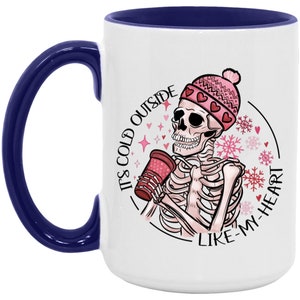 May include: White ceramic mug with a navy blue handle and interior. The mug features a skeleton illustration wearing a pink hat and holding a red cup. Text on the mug reads "It's cold outside like my heart."