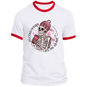 May include: White ringer t-shirt with red trim featuring a skeleton wearing a pink beanie and holding a coffee cup. The graphic includes the text "IT'S COLD OUTSIDE LIKE-MY-HEART" in a circular design with snowflakes.