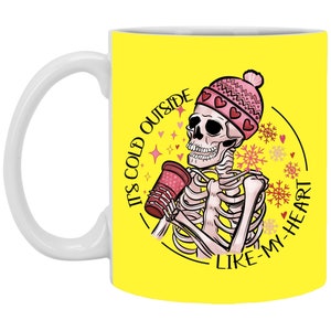 May include: White ceramic mug with a yellow background featuring a skeleton wearing a pink hat with hearts, holding a red cup. The mug has the text "IT'S COLD OUTSIDE LIKE-MY-HEART".